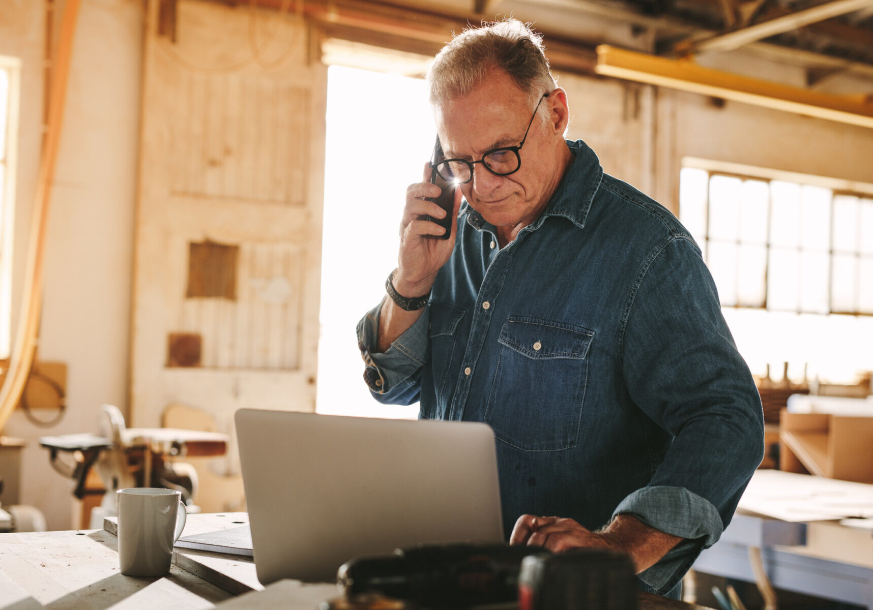 Senior man talking on cell phone and using laptop on work table. Mature carpenter working on laptop and answering phone call in his carpentry workshop.